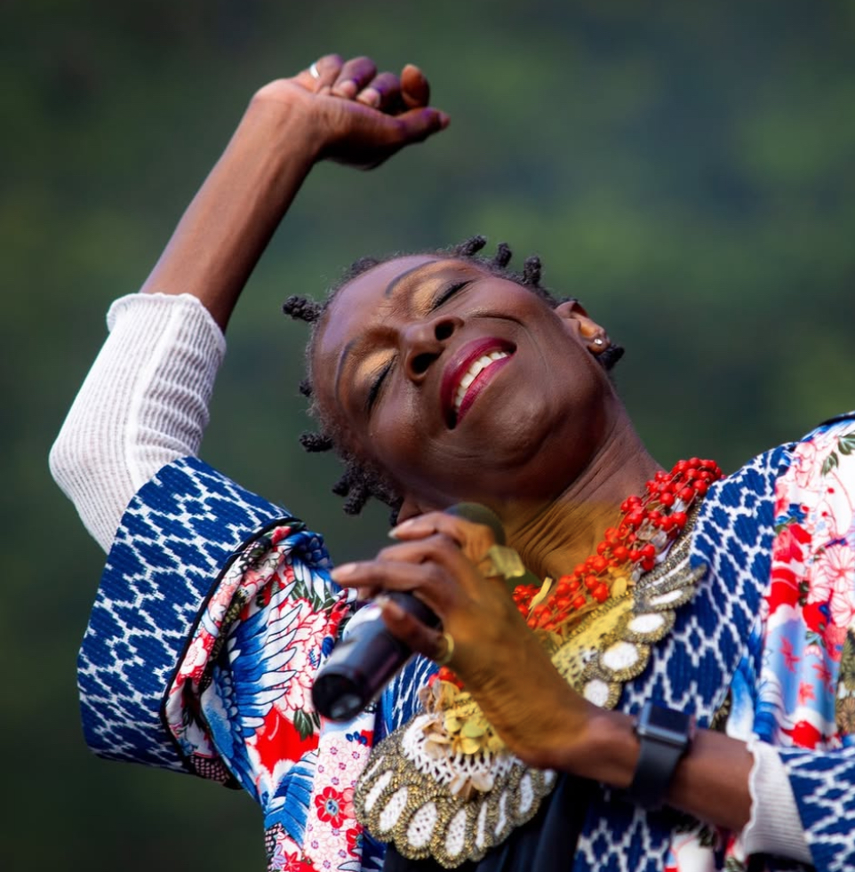 Performer singing with arm raised, eyes closed, wearing a colourful patterned outfit and layered beaded necklaces