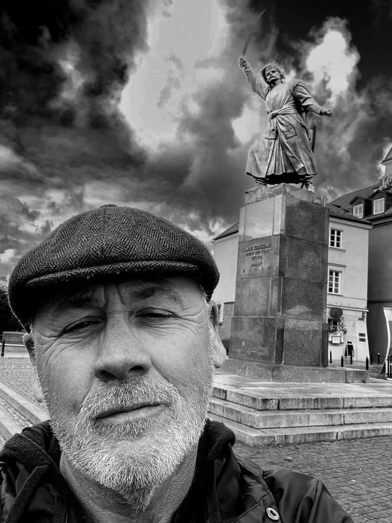 Black and white self-portrait of the photographer wearing a flat cap, with a statue and dramatic sky in the background